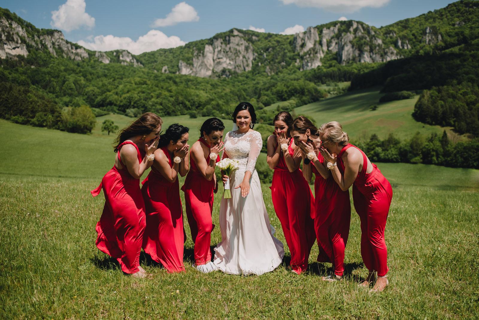 6 družičiek na svadbu v červených šatách na lúke. Družičky obdivujú zásnubný prsteň.